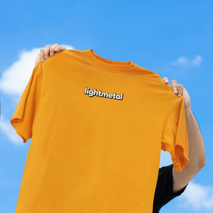 Wild Yellow - Person holding an orange t-shirt with 'lightmetal' logo against a blue sky.