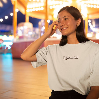 Corporate Beige - Woman wearing a light gray t-shirt with 'lightmetal' logo in an outdoor setting with blurred lights in the background