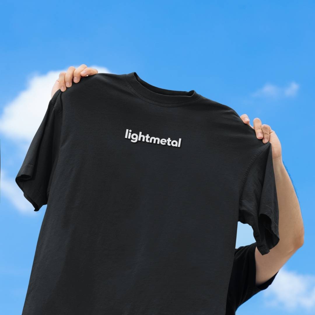 Honest Black - Person holding a black t-shirt with 'lightmetal' logo against a blue sky.